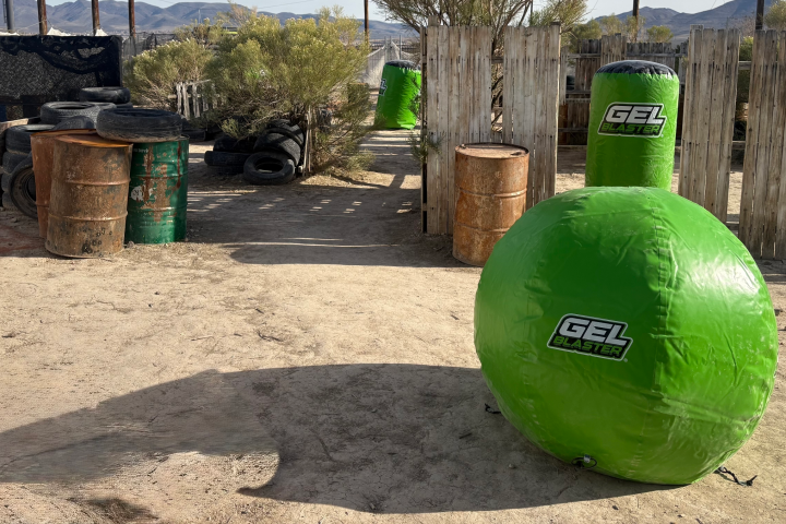 Outdoor game area with inflatable green barriers labeled 'Gel Blaster' and rustic fences and barrels.