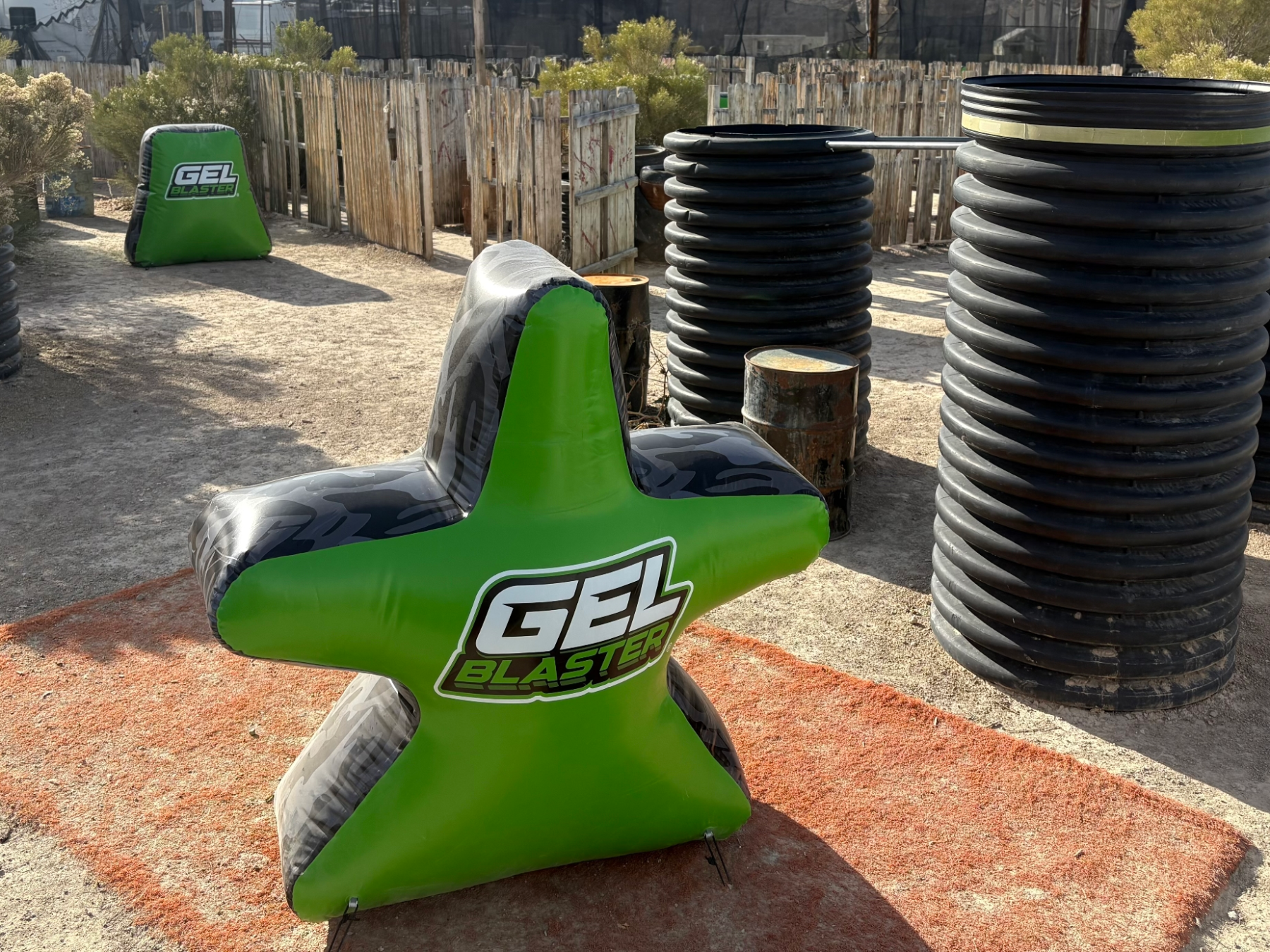 Outdoor paintball field with inflatable green star barriers labeled 'Gel Blaster' and cylindrical cover.