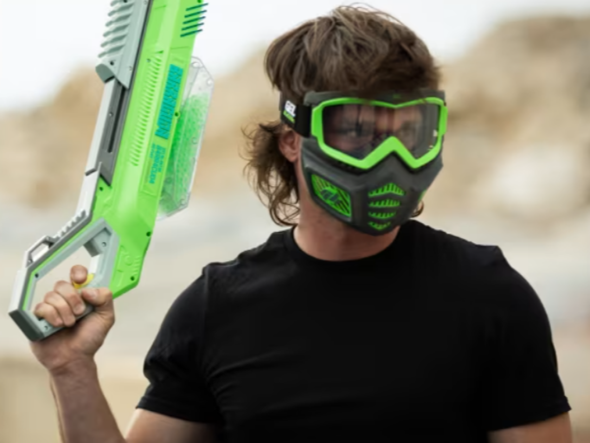 Person in black shirt wearing green mask holds a large green toy gun.