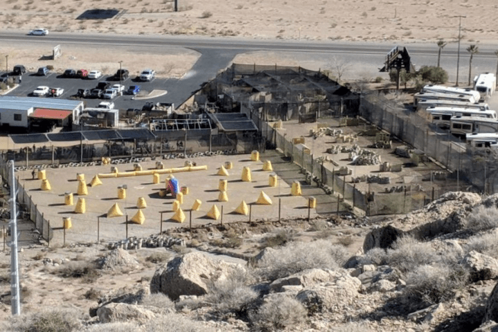 Aerial view of desert paintball field with yellow obstacles and nearby parked cars and trailers.
