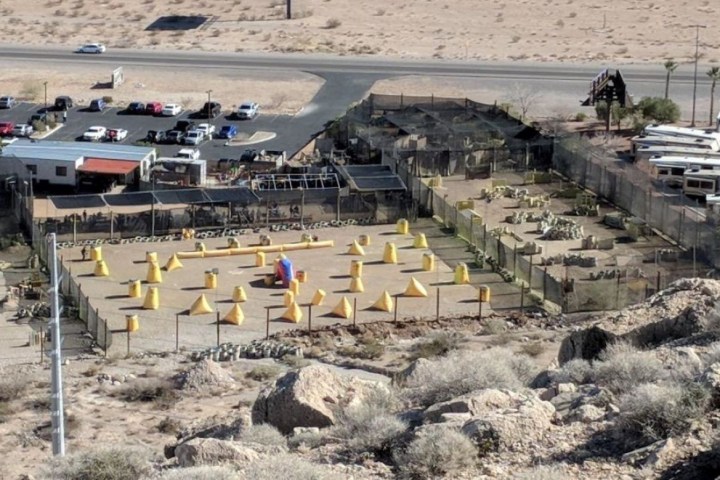 Aerial view of a fenced paintball field with yellow and red obstacles in a desert area.