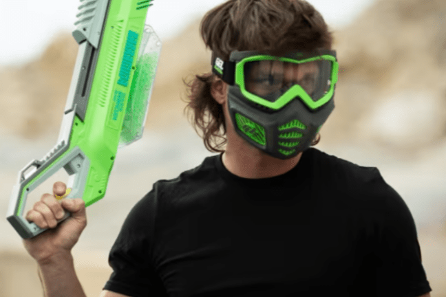 Person wearing a green mask and holding a large green toy gun, standing outdoors.