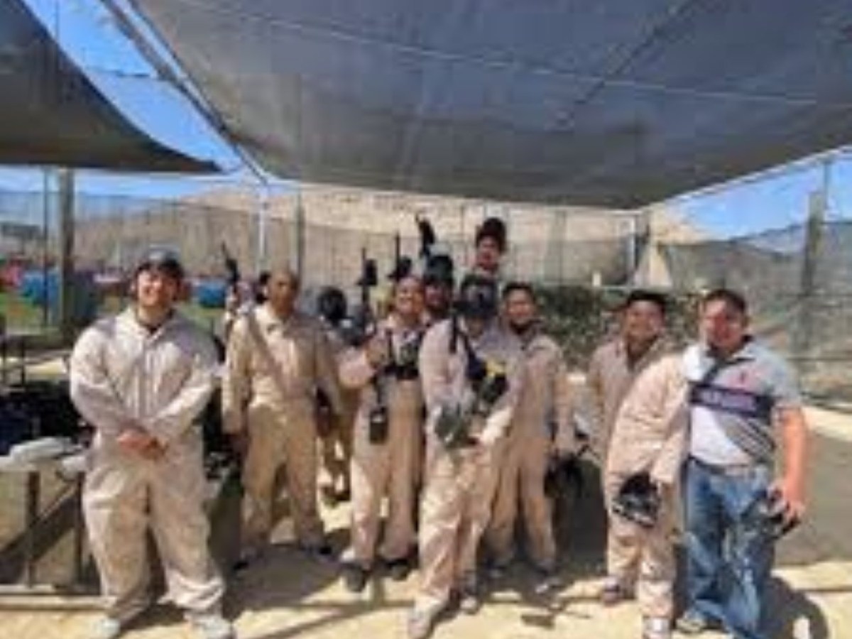 Group of people in beige jumpsuits holding paintball gear under a canopy outdoors.