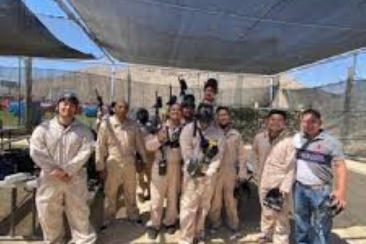 Group of people in beige jumpsuits holding paintball gear under a canopy outdoors.