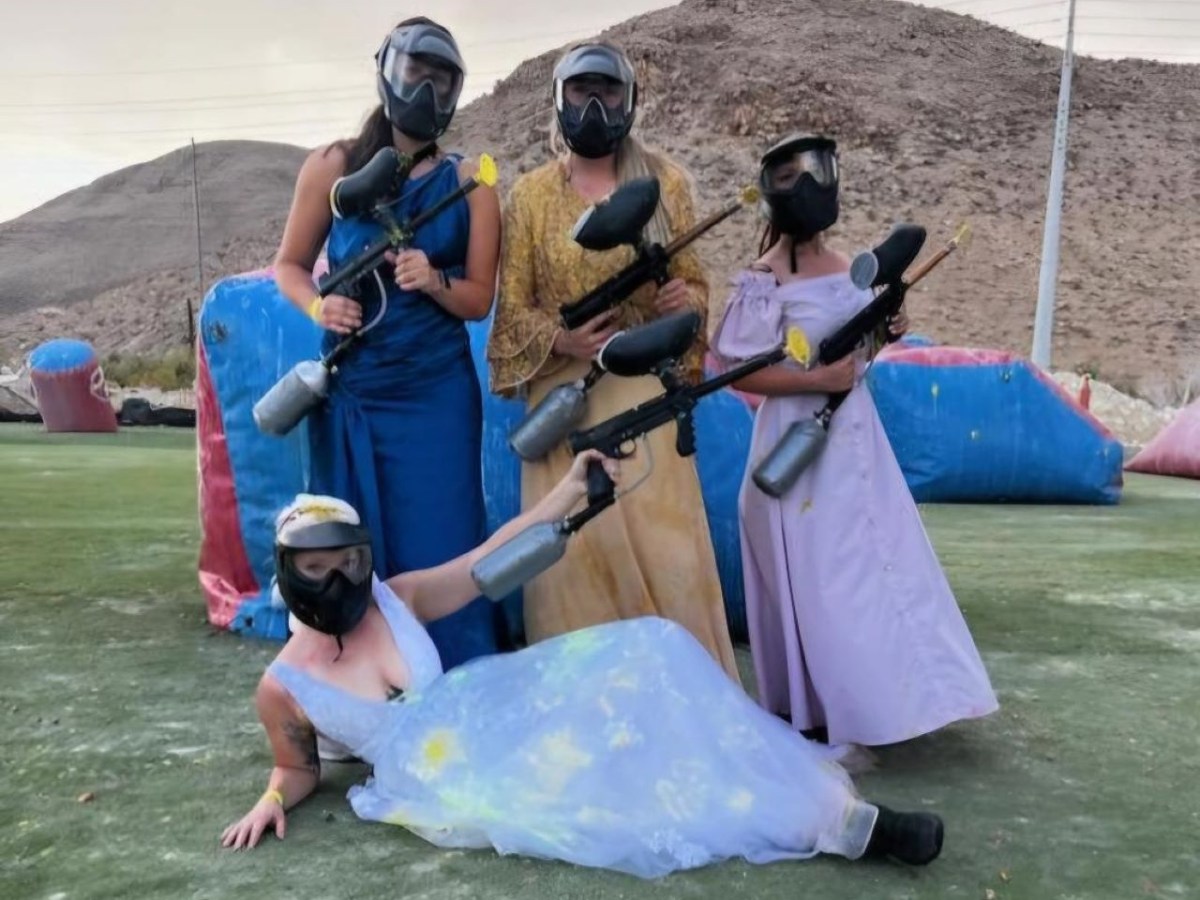 Four people in dresses and masks holding paintball guns on a field.