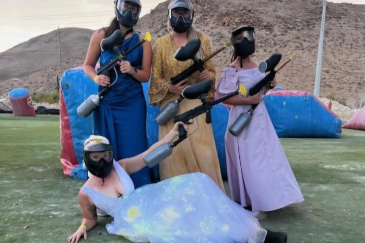 Four people in dresses and masks holding paintball guns on a field.