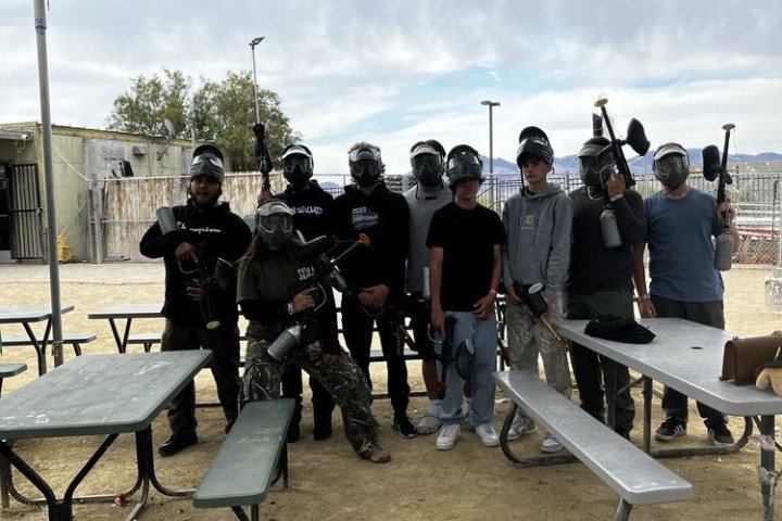 Group of people in paintball gear posing behind picnic tables outdoors.