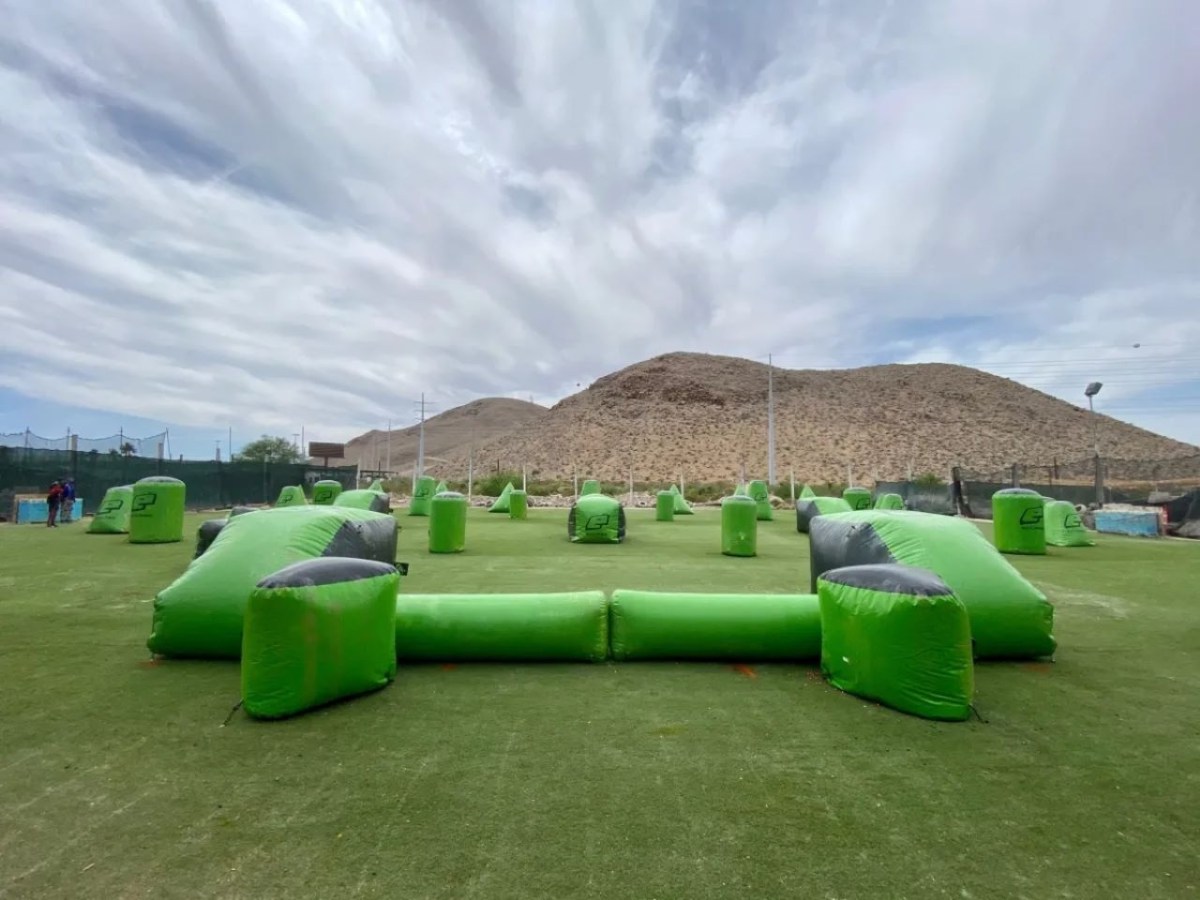 Paintball field with green inflatable obstacles on grass, desert hills in background.