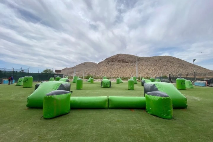 Paintball field with green inflatable obstacles on grass, desert hills in background.
