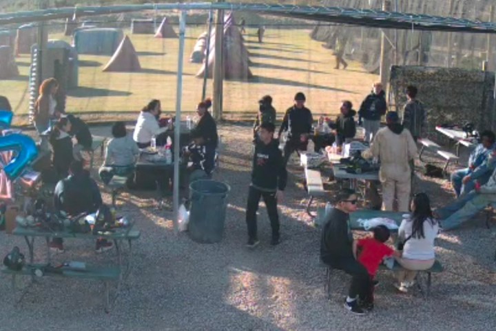 Group of people gathered outdoors with tables and paintball field in background, blue balloon in foreground.