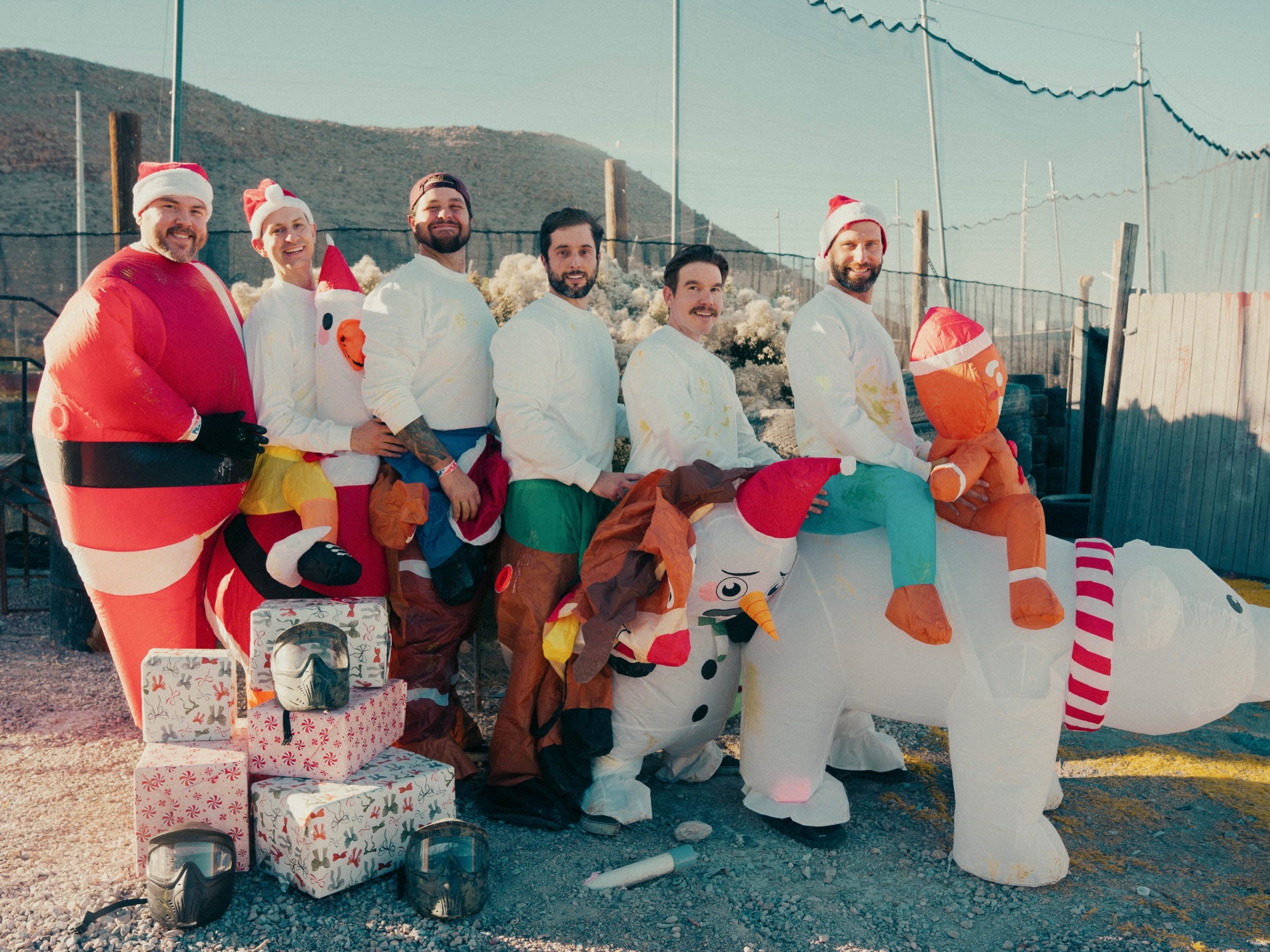 Group of six people wearing inflatable Christmas costumes with gift boxes outdoors on a sunny day.