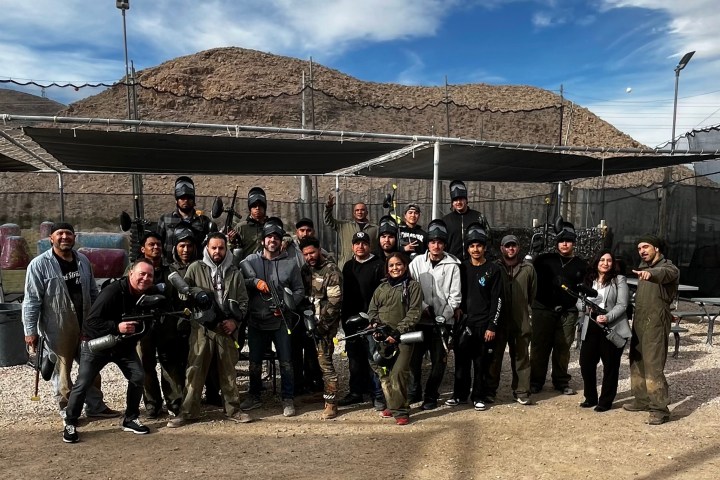 Group of people in paintball gear posing outdoors with mountains in the background.