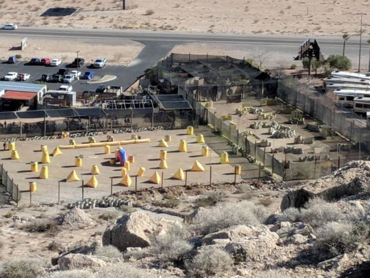 Aerial view of outdoor paintball field with yellow obstacles, near a road and parked vehicles.