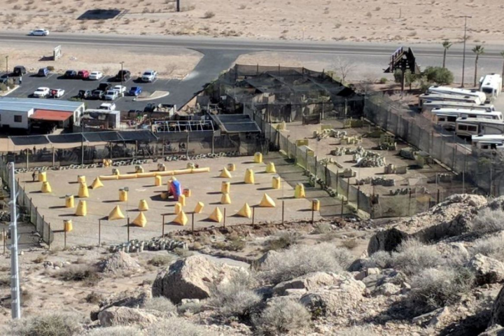 Aerial view of outdoor paintball field with yellow obstacles, near a road and parked vehicles.