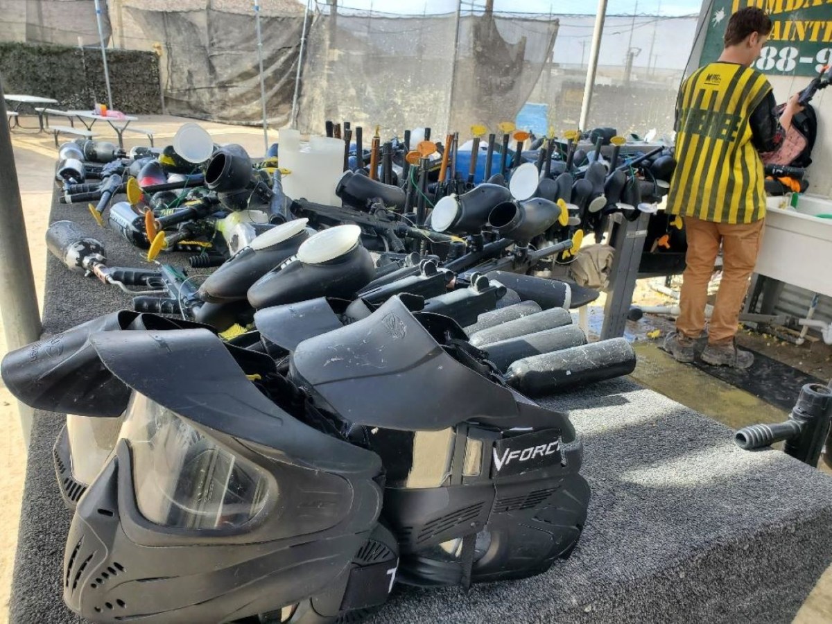 Paintball masks and guns on a table, person cleaning equipment in the background.