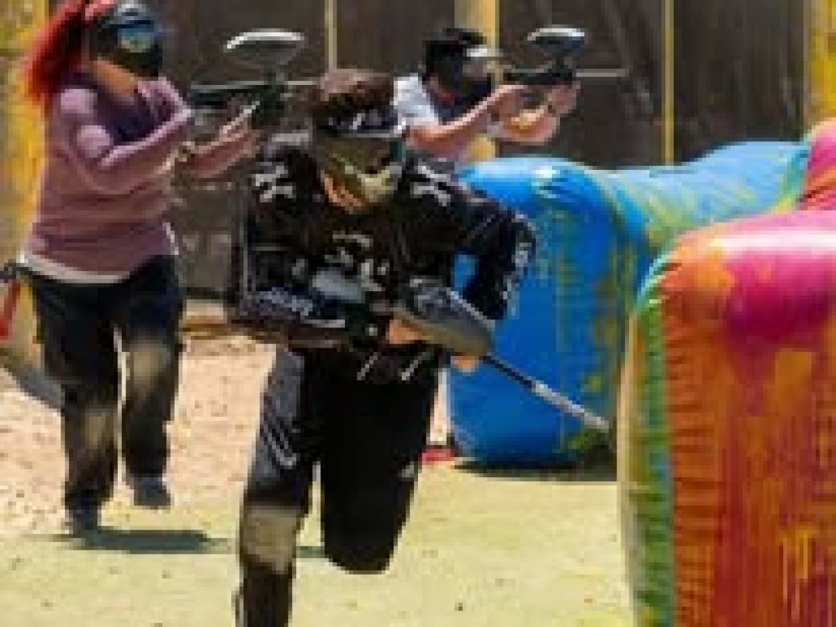 Three people play paintball, running and aiming markers behind inflatable obstacles.