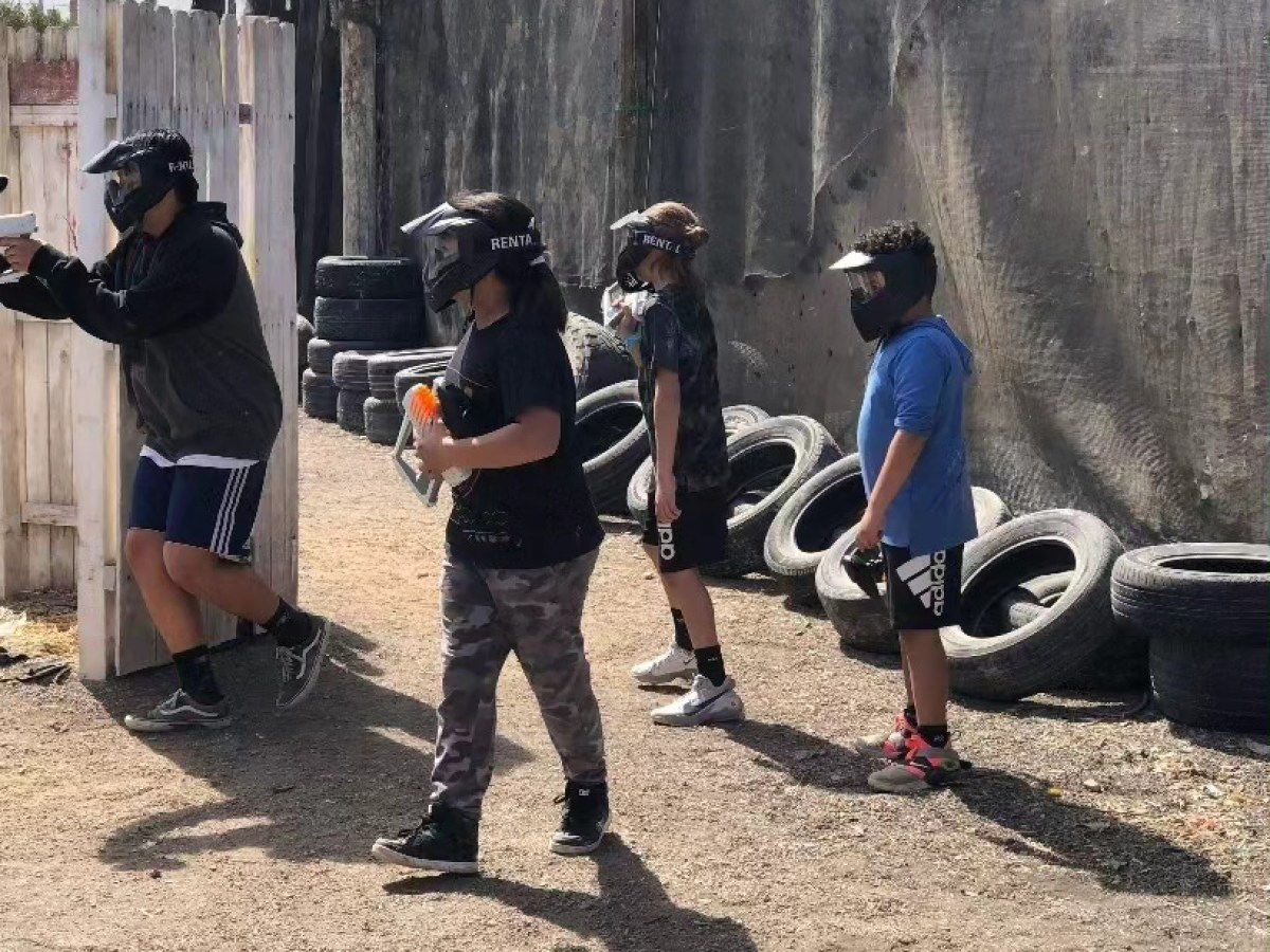 Four kids wearing helmets holding toy guns in an outdoor play area with tires.