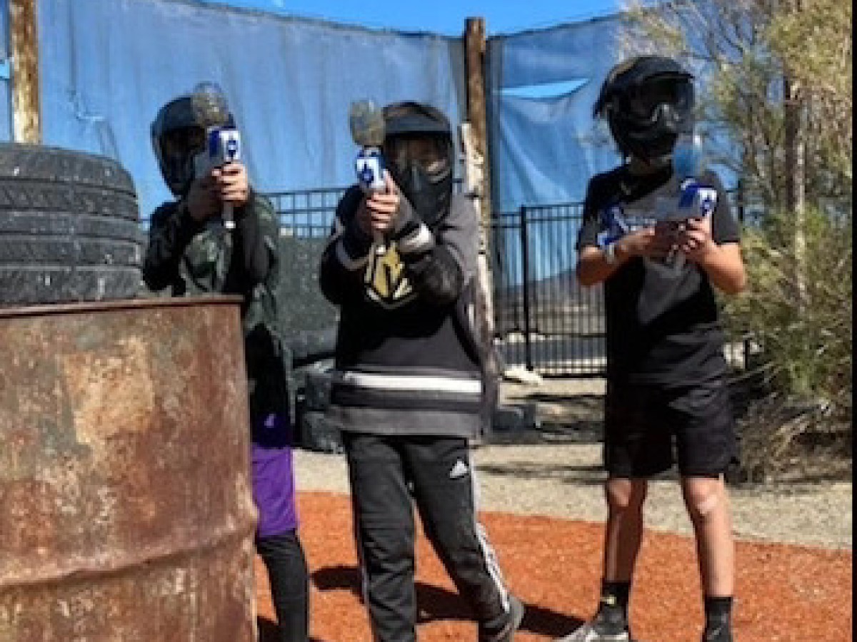 Three kids in paintball gear aiming markers outdoors by a rusty barrel and stacked tires.