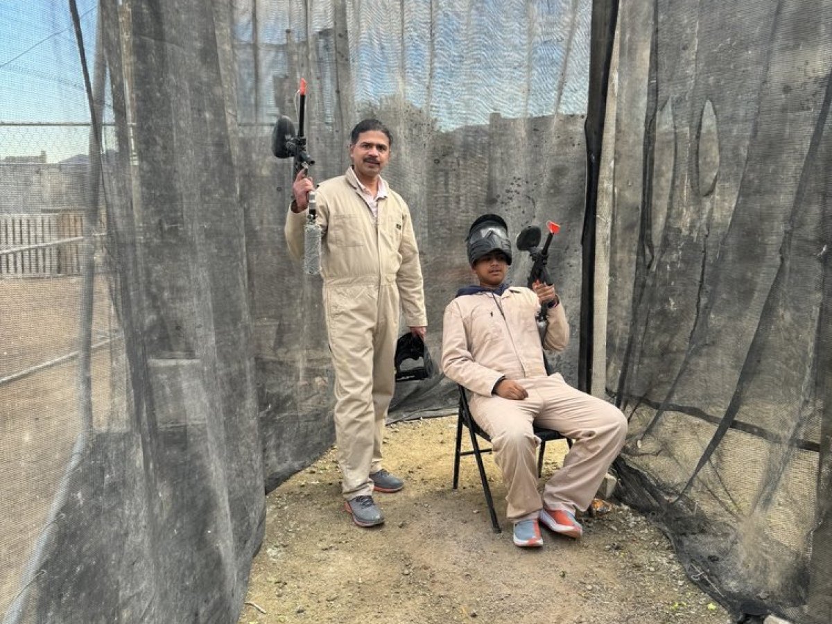Two people in beige jumpsuits holding paintball guns, one standing, one sitting in a chair inside a mesh enclosure.
