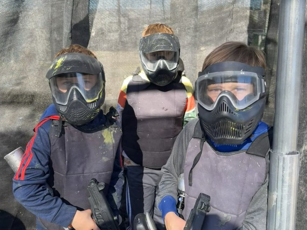 Three kids in protective gear holding paintball guns, standing together outdoors.