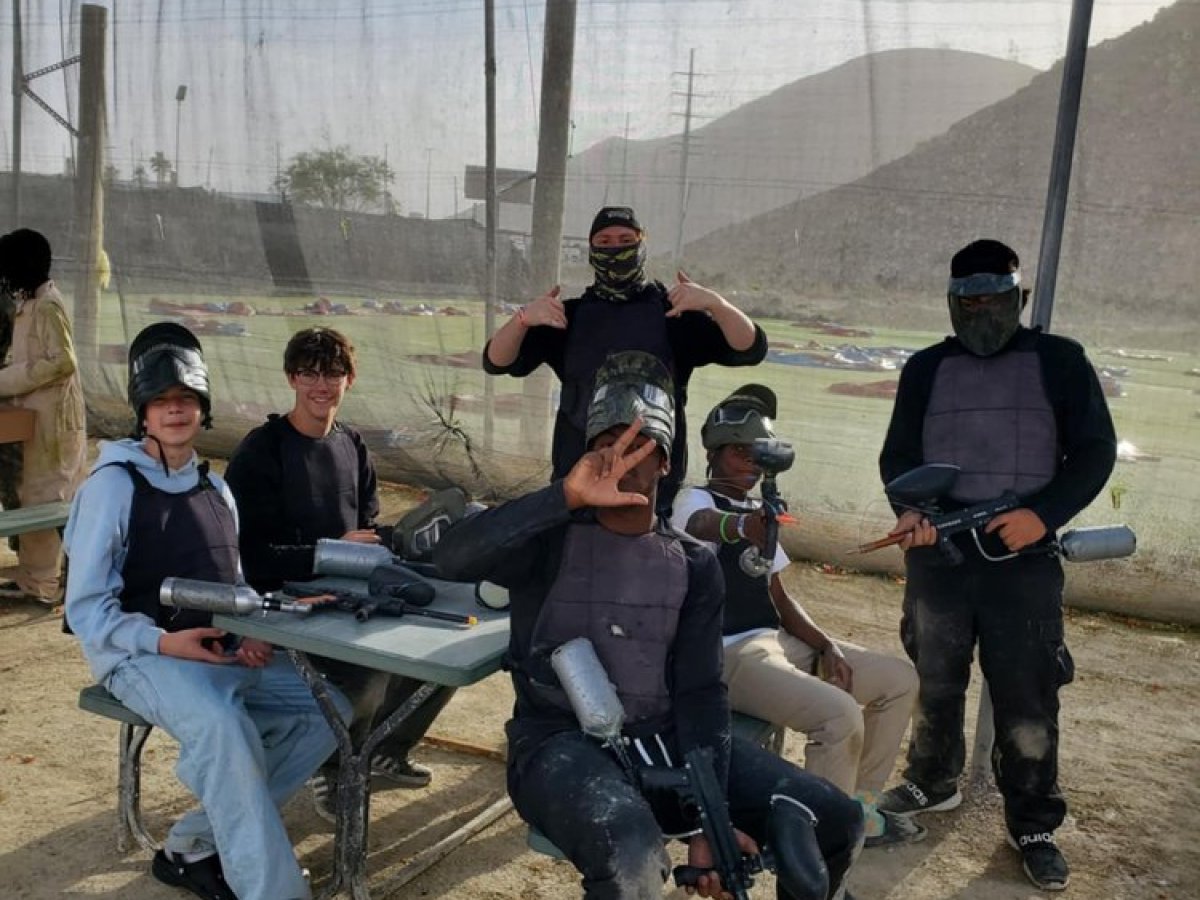 Group of six people in paintball gear posing outdoors, some sitting on a bench.