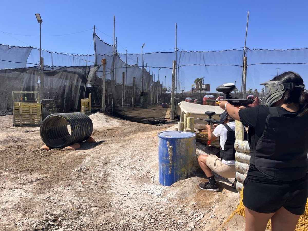 Two people playing paintball, crouching behind barriers on a sunny day.